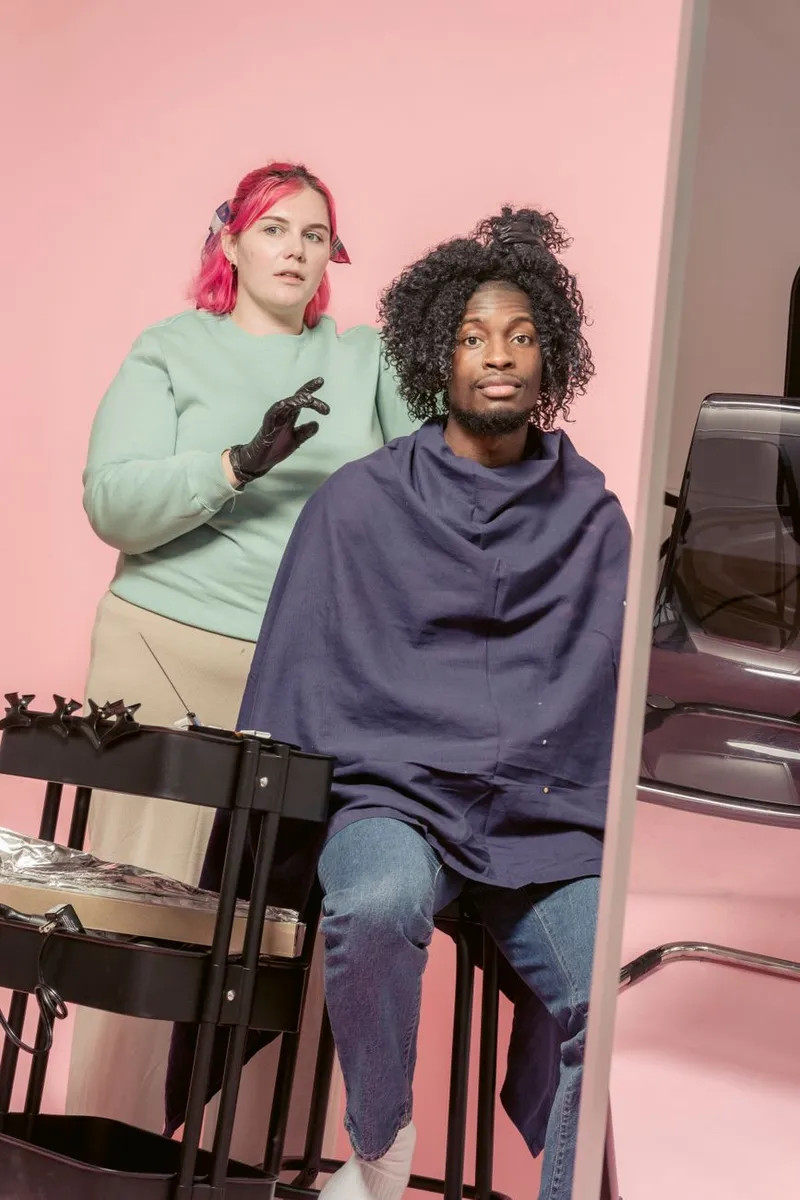 Female hairstylist with bright dyed hair doing hairstyle to African American customer wrapped in cape in studio against pink background